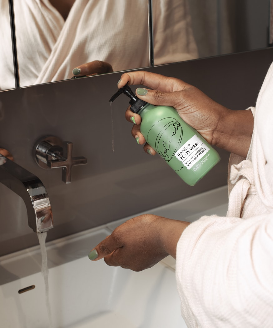 photo in a bathroom with grey wiles and a white sink with chrome tap. Close up of a black woman's torso and hands as she hold the upcircle beauty kiwi and lemongrass hand and body wash in her hands as if she is going to dispense some to wash her hands. she is wearing a white cloth robe and green nail varnish and you can see her torso reflected in the mirror