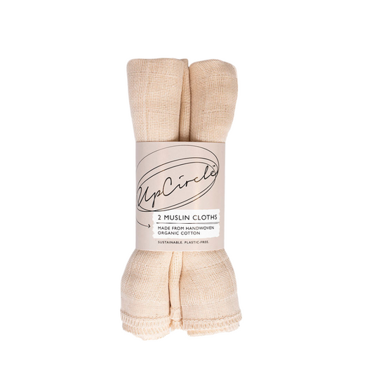 Two rolled muslin cloths are wrapped with a label that reads "UpCircle." The label indicates that the cloths are handwoven from organic cotton and packaged sustainably. The cloths are beige in colour and have a simple, textured appearance.