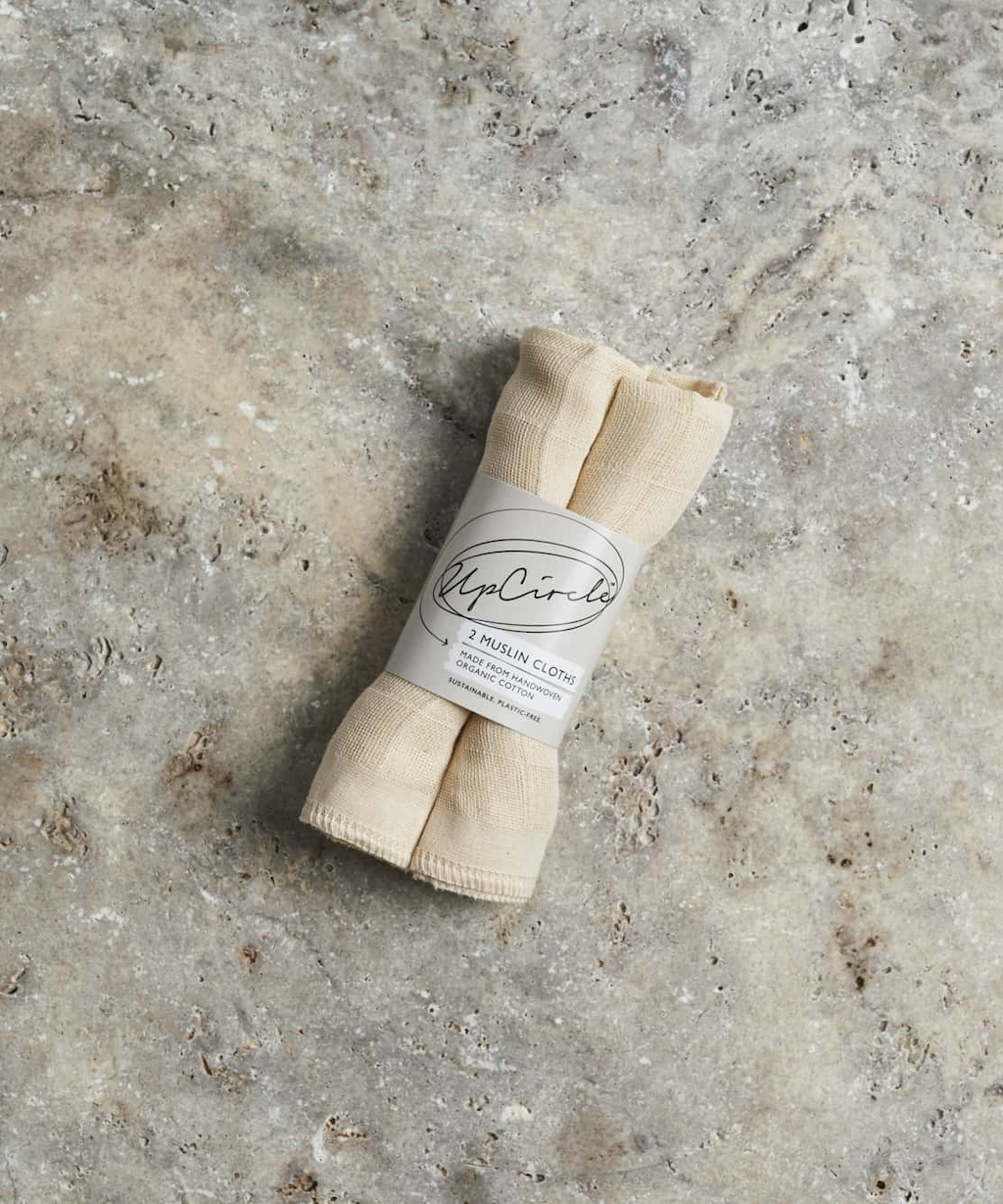 2 neatly rolled beige muslin cloths are wrapped in a paper label that reads "UpCircle 2 Muslin Cloths.” The cloth is placed on a textured grey and brown mottled surface.