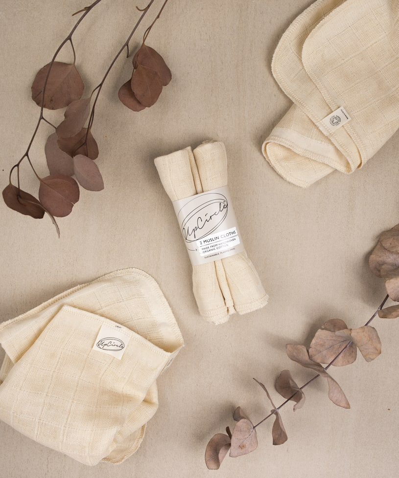 Beige reusable cloths, some folded and some loosely arranged, are displayed on a light brown surface. Dried eucalyptus branches frame the image, adding a natural element. The cloths feature a small logo tag that reads upcircle.