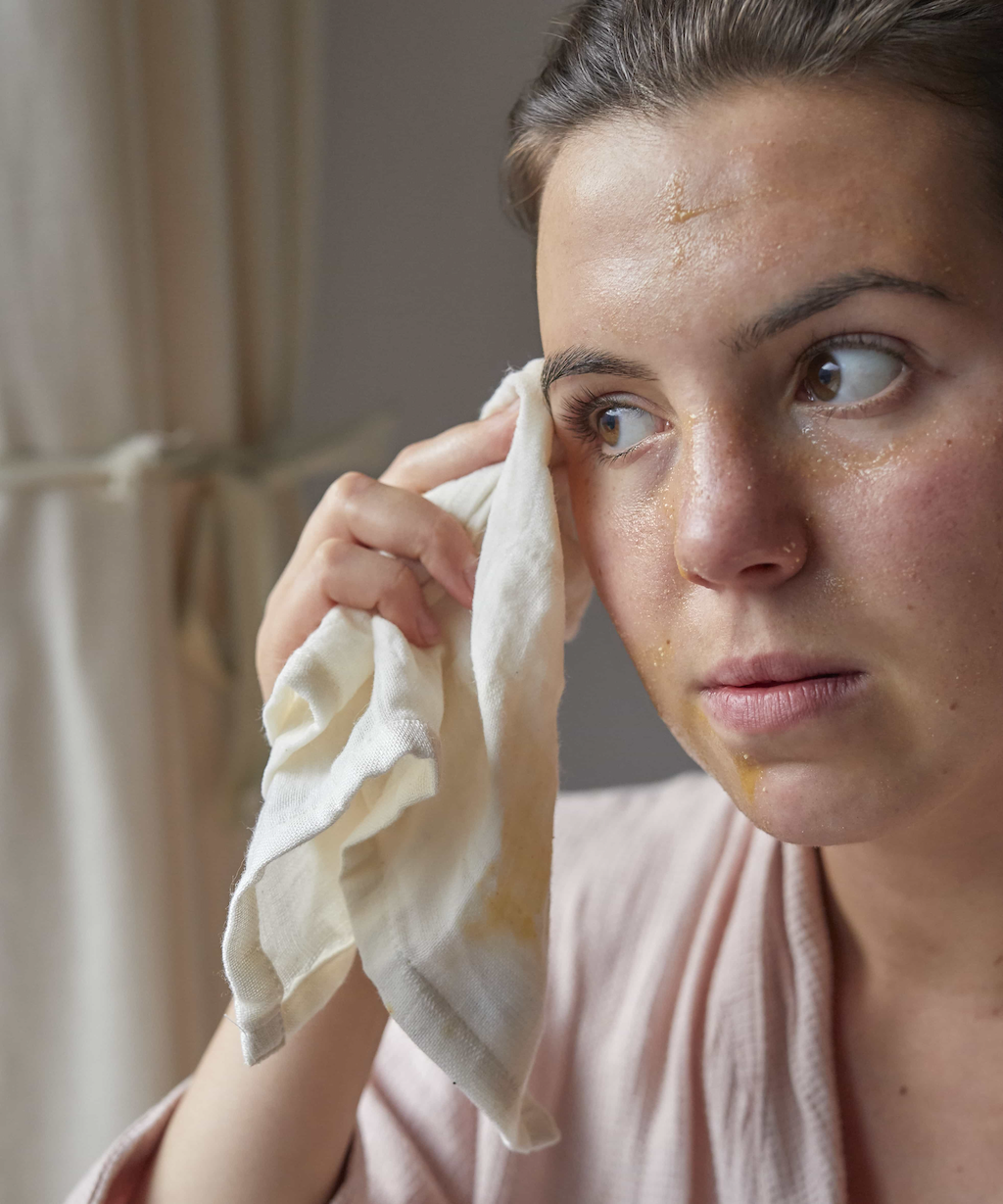 A person with a focused expression wipes their face with a white cloth while appearing to remove a facial mask or treatment. They have light brown hair pulled back and are wearing a light pink bathrobe. A window with light-colored curtains is visible in the background.