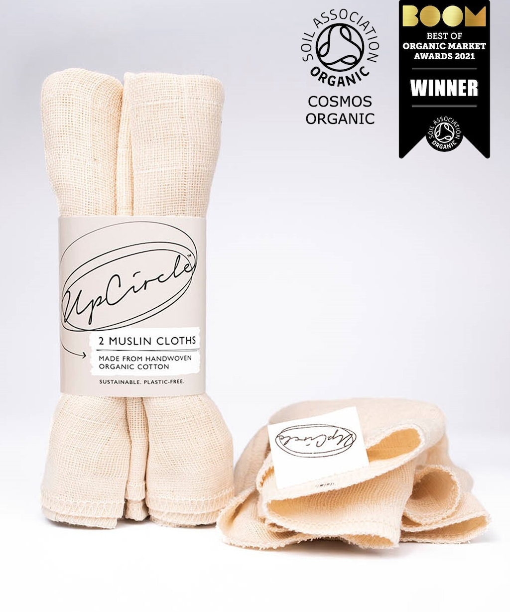 The image shows two rolls of handwoven organic cotton muslin cloths from UpCircle, packaged with a label. One roll stands upright while the other is laid flat and partially unwrapped. Award logos for "Soil Association," "Cosmos Organic," and "BOOM 2021 Winner" are displayed.