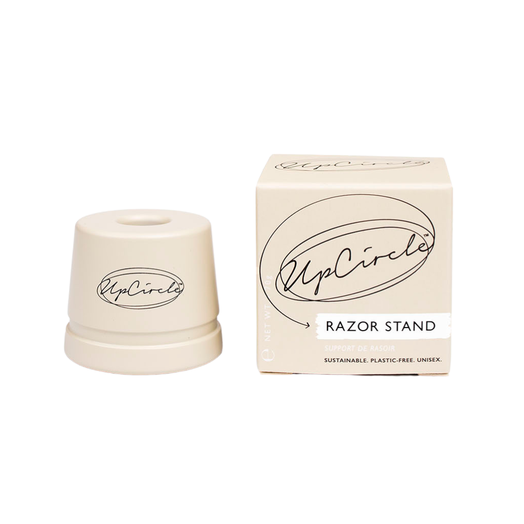 A circular, cream-coloured razor stand is stood against a white background and placed next to its packaging; a cream-coloured cube with the product and brand name listed in black.