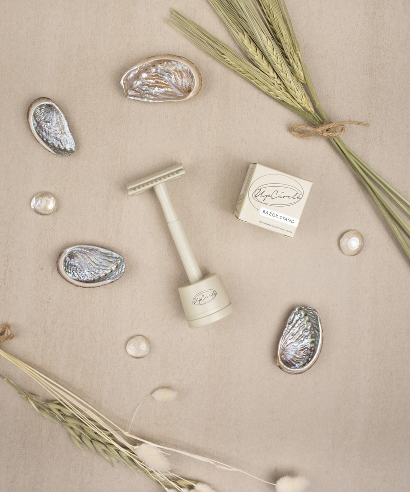 Top view of a beige background displaying a white razor on a stand with a matching box labeled "Razor Stand." The scene is decorated with seashells, wheat stalks, and round white stones, creating a natural and earthy aesthetic.