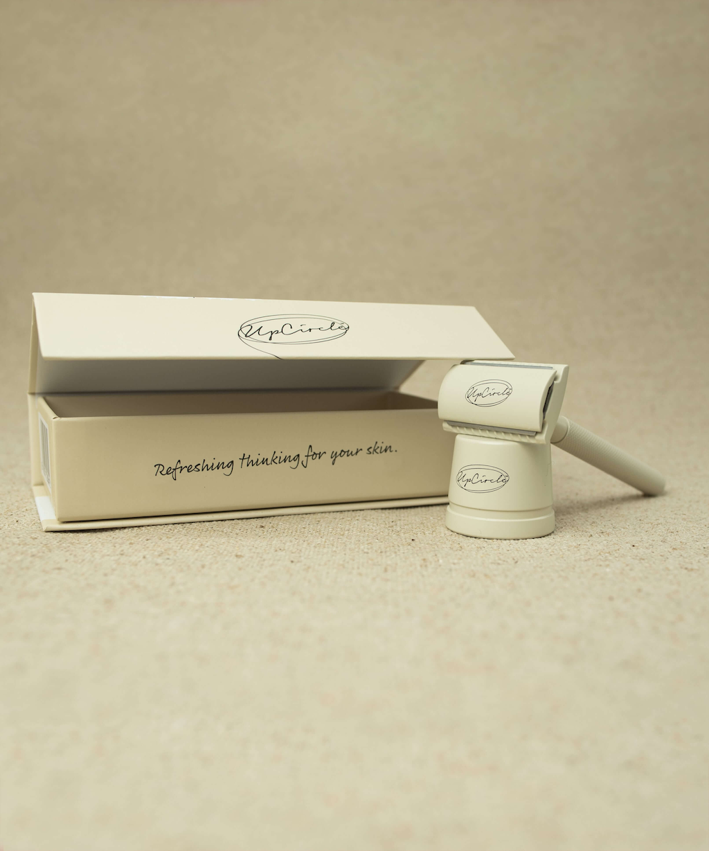 The safety razor positioned standing up next to its matching box. The box displays the brand logo "UpCircle" and the slogan "Refreshing thinking for your skin." The background is a neutral beige color and sandy texture