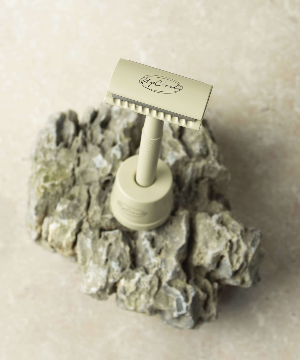 Upcircle's vintage-style safety razor with a textured handle, standing upright on a rough, gray stone surface. The razor is engraved with a logo and has a classic, minimalist design. The background is a soft, light-colored textured surface.
