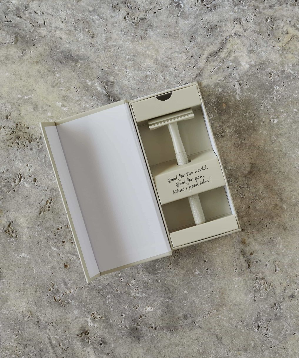 A cream, minimalist safety razor in a beige box, placed on a textured stone surface. The box lid is open, revealing the razor and an inner note that reads, "Good for the world, great for your wallet, best choice!" in handwritten script.