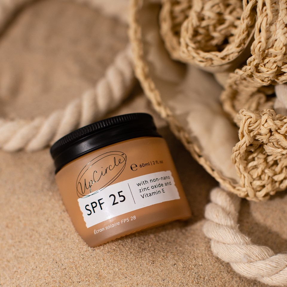 A small jar of UpCircle SPF 25 sunscreen with non-nano zinc oxide and Vitamin E rests on sandy ground. A woven straw-like bag and thick rope are nearby, adding a beachy, summer vibe to the scene.