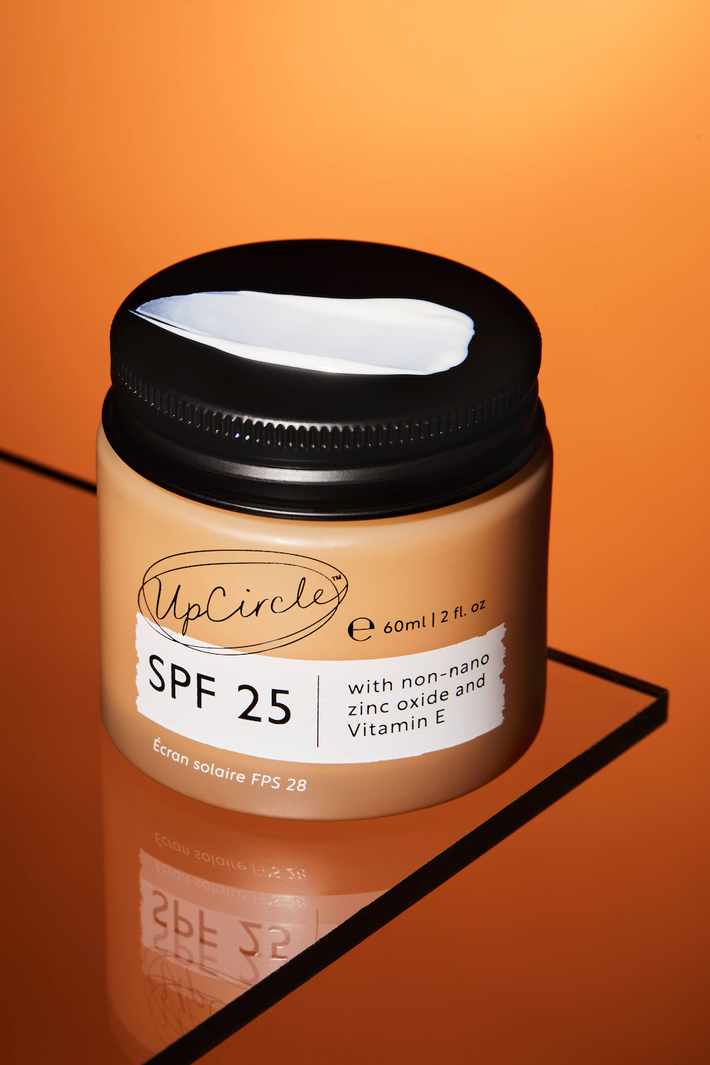 A jar of UpCircle SPF 25 sunscreen with non-nano zinc oxide and Vitamin E. The jar is 60 ml (2 fl oz) and features a black lid. The background is an orange hue, and the jar is placed on a reflective surface with a black border.