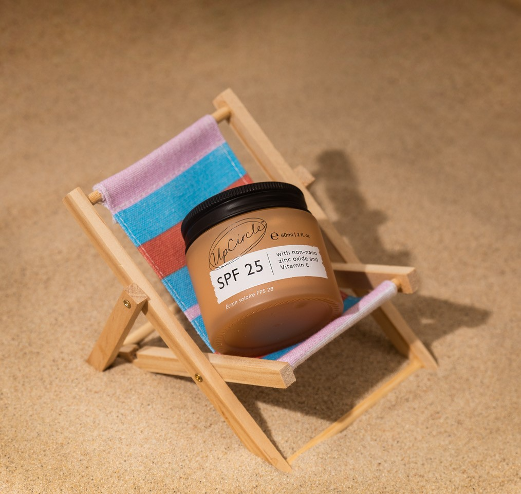A jar of SPF 25 sunscreen lotion from UpCircle is placed on a small wooden beach chair with a striped fabric seat. The scene is set on sandy ground, reminiscent of a beach setting. The label on the jar mentions it contains zinc oxide and vitamin E.