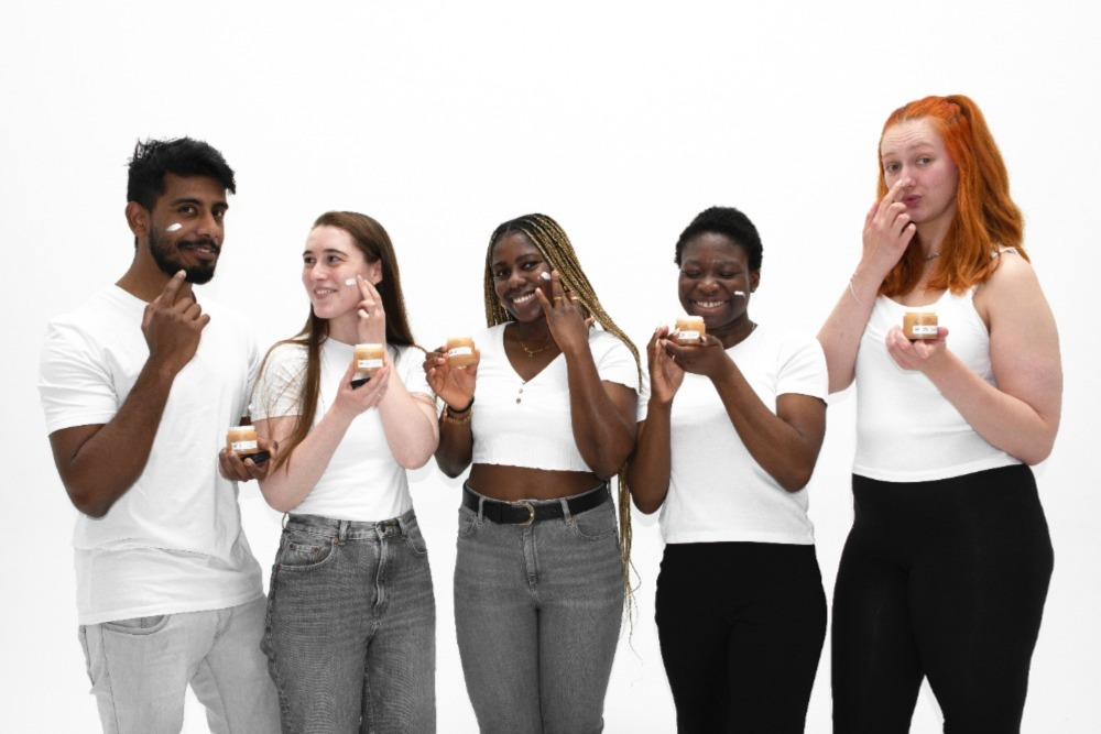 A diverse group of five people stands in a row against a plain white background, each holding a small jar of cream. They are all wearing casual white tops and smiling, while some apply the cream to their faces.