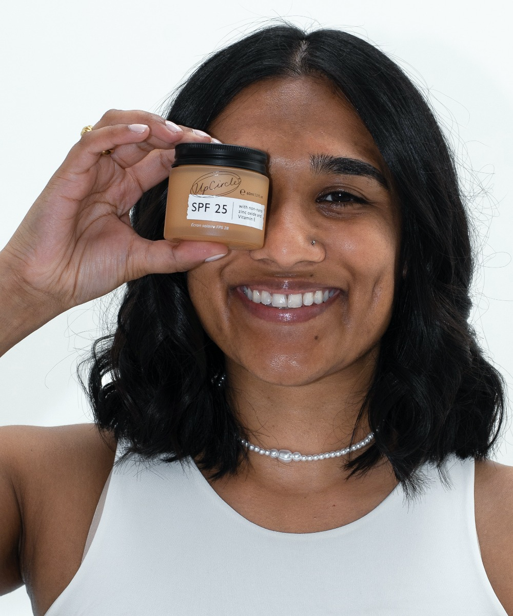 A person with shoulder-length dark hair holds a jar of SPF 25 sunscreen in front of one eye and smiles at the camera. They are wearing a white top and a beaded necklace. The background is plain white.