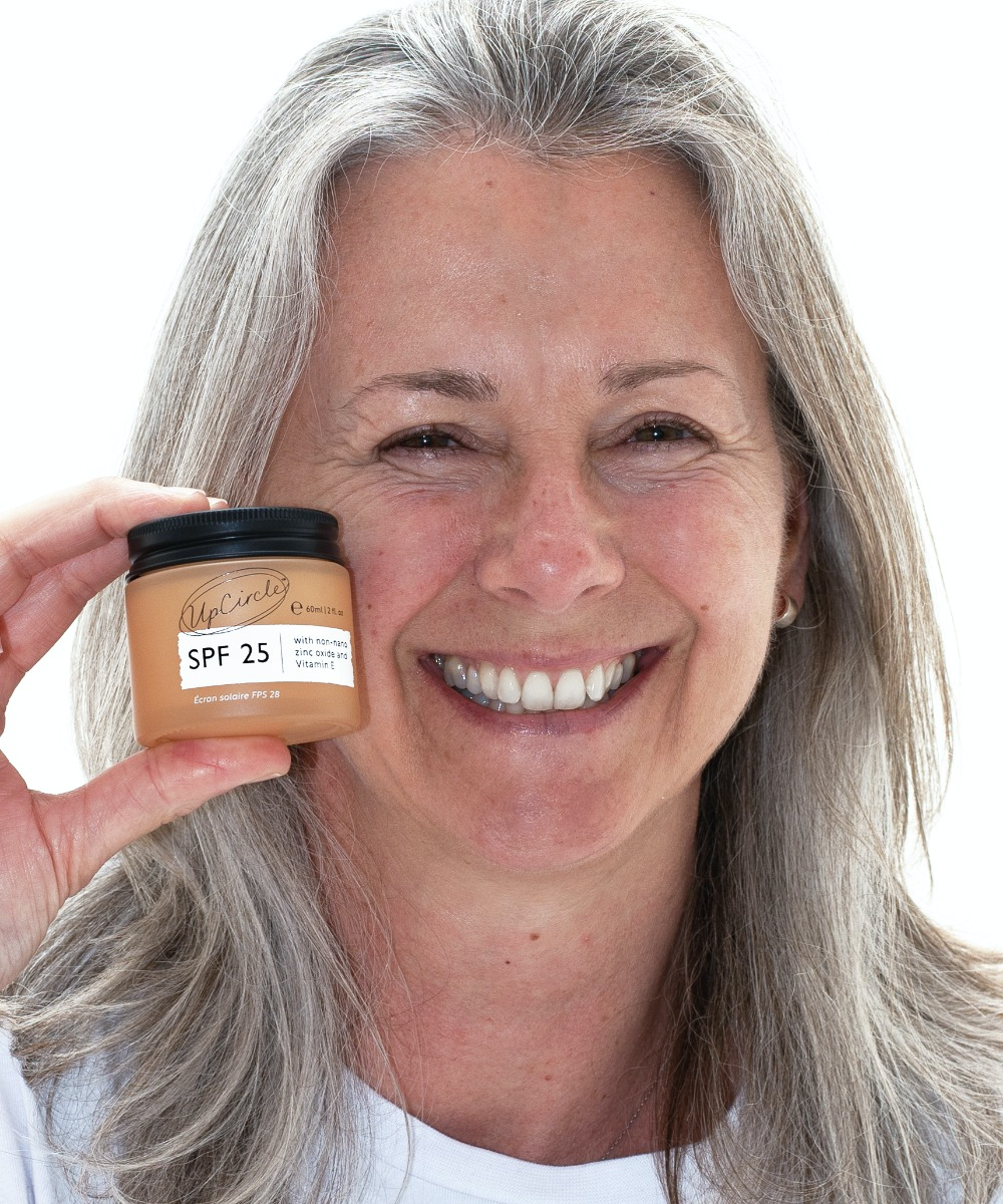 A smiling woman with gray hair holds a jar of UpCircle SPF 25 sunscreen close to her face. She is wearing a white top and standing against a plain white background. The sunscreen jar has a beige label with text indicating its SPF protection.