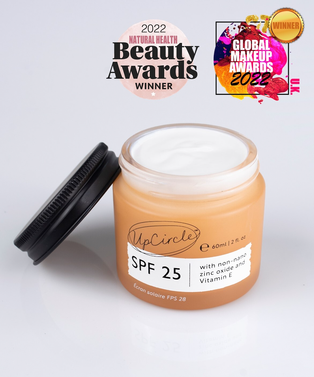 A 60ml jar of UpCircle SPF 25 cream with a black lid is open, revealing creamy white contents. The jar label mentions non-nano zinc oxide and Vitamin E. Two award stickers are visible: "2022 Natural Health Beauty Awards Winner" and "Global Makeup Awards 2022 Winner.