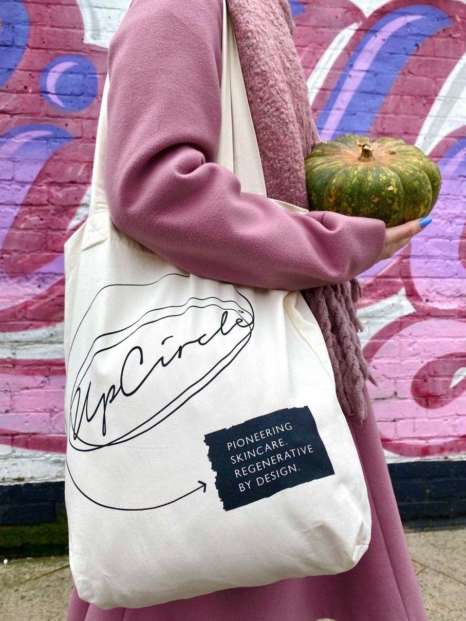 A person in a pink outfit holds a green pumpkin and wears a white tote bag with the UpCircle logo. The tote bag has the text: "Pioneering Skincare. Regenerative by Design." The background features a pink and purple graffiti-covered brick wall.