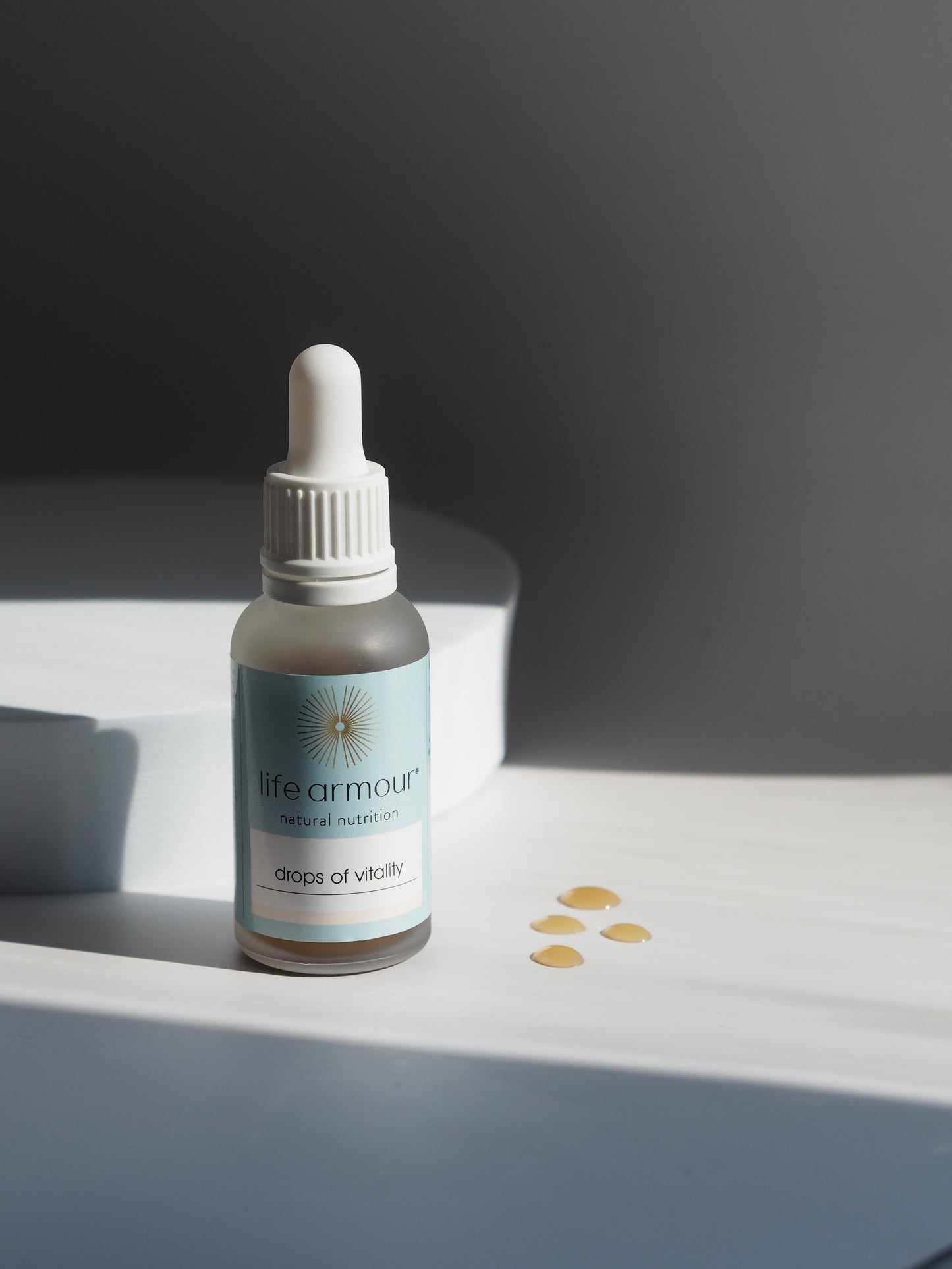 A small dropper bottle with a white cap labeled "life armour natural nutrition drops of vitality" sits on a surface with four drops of liquid nearby. The bottle is illuminated by soft light, casting gentle shadows in a minimalist setting.