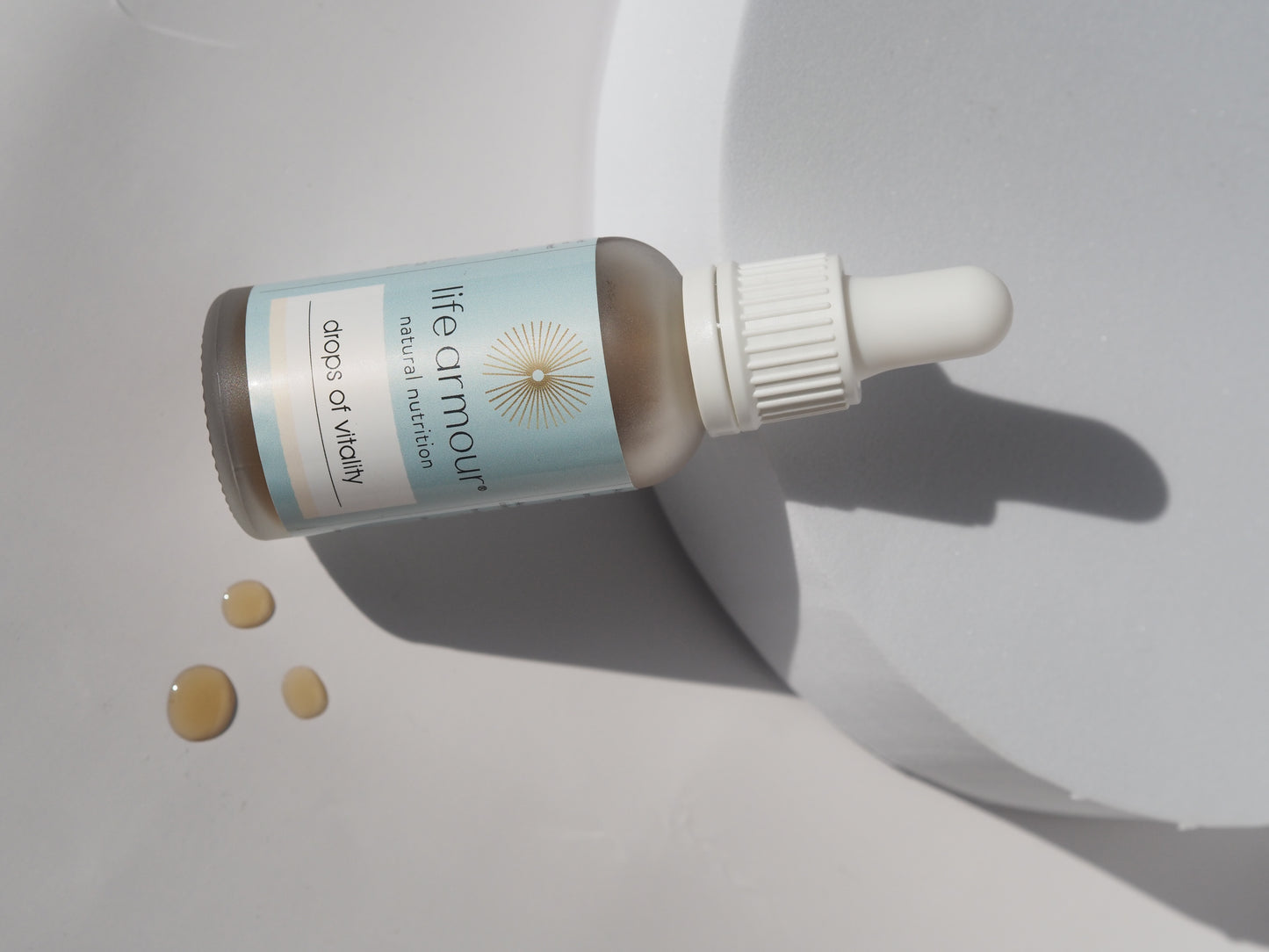 A bottle of Life Armour "Drops of Vitality" tincture lies on its side on a smooth, light grey surface. The bottle is pastel blue with a white dropper cap and some liquid drops are spilled beside it. The product casts a soft shadow.