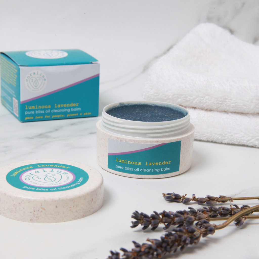 A jar of "Luminous Lavender" pure bliss oil cleansing balm sits open on a marble surface, with its lid beside it. A box of the product, a white towel, and dried lavender sprigs are in the background. The balm has a smooth lavender color and a serene setting.