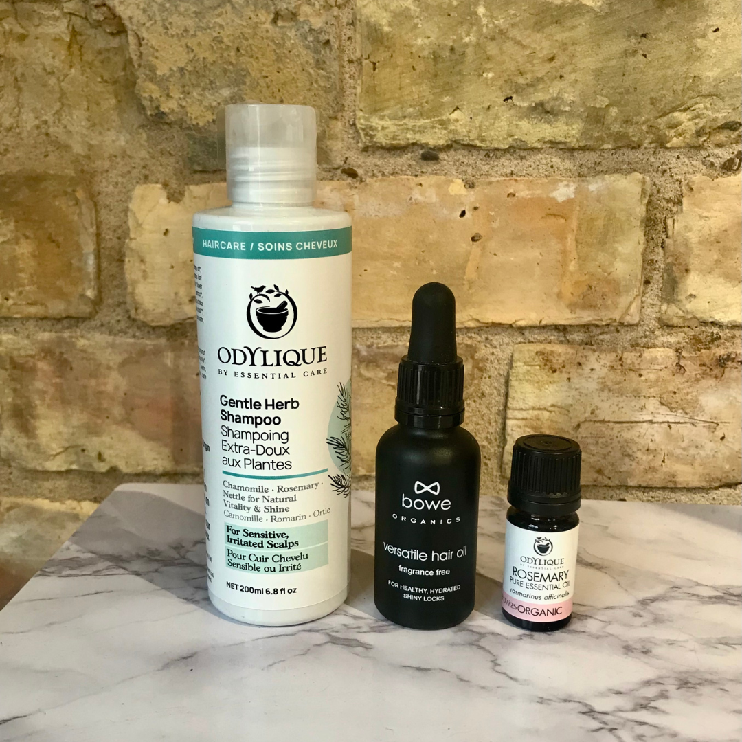 3 products that are good for hair and scalp health and growth are photographed on marble effect table and exposed brick background. Left to right odylique's gentle herb shampoo, bowe organics fragrance free versatile hair and oil and odylique's organic rosemary essential oil