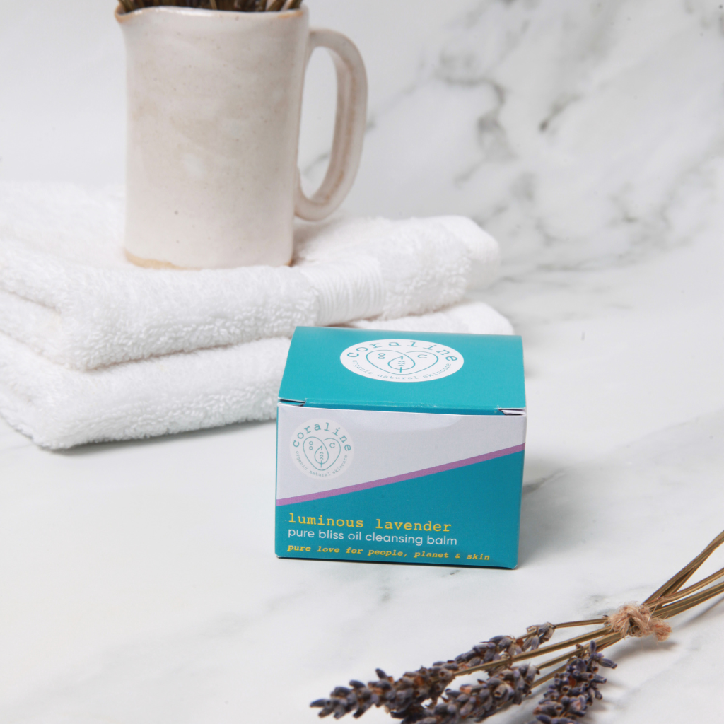A small turquoise box of "Luminous Lavender Pure Bliss Oil Cleansing Balm" from Coraline Skincare placed on a marble surface. In the background, there is a ceramic mug with lavender sprigs, a stack of white towels, and additional lavender sprigs in the foreground.