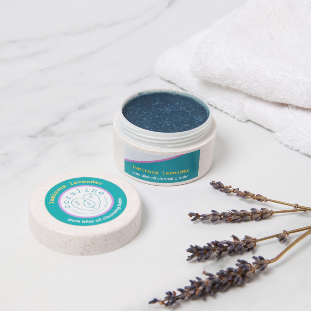 A jar of "Luminous Lavender - pure bliss oil cleansing balm" with its lid off is placed on a white marble surface. A sprig of lavender and two folded white towels are beside it. The product's box is in the background.