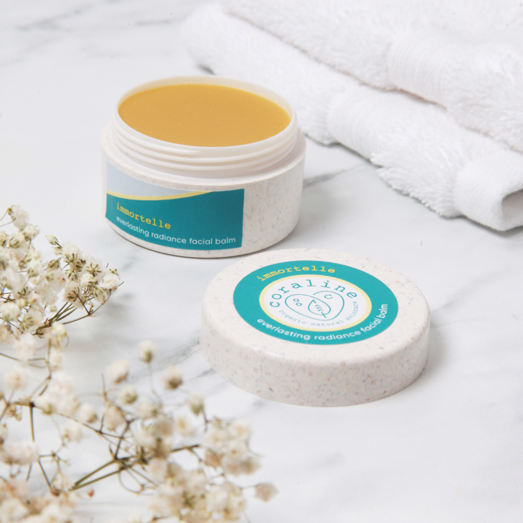 lifestyle image of the immortelle facial balm which can be seen on a white stone surface with some white cotton fluffy flannel cloths behind the open bio plastic pot of the balm. inside the pot you can see the orange colour of the face balm. in the foreground are some dried gypsophilia flowers