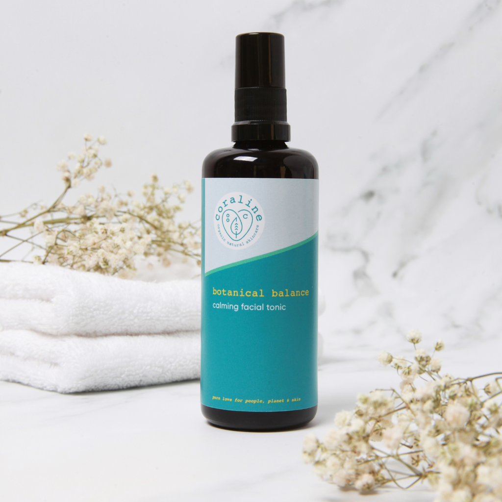 coraline skincare botanical balance calming facial tonic is in a black bottle with a blue label and is in a white and grey marble background with white towels and some dried cream flowers