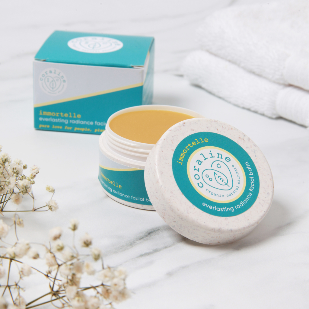 lifestyle photo of the coraline immortelle face balm. the product is on a white background with fluffy white towels and dried white bud flowers. in the background you can see the sea blue box packaging for the balm and the white pot for the balm in the foreground with the jar opened and the lid resting on the side of the jar. the balm inside is an orange colour