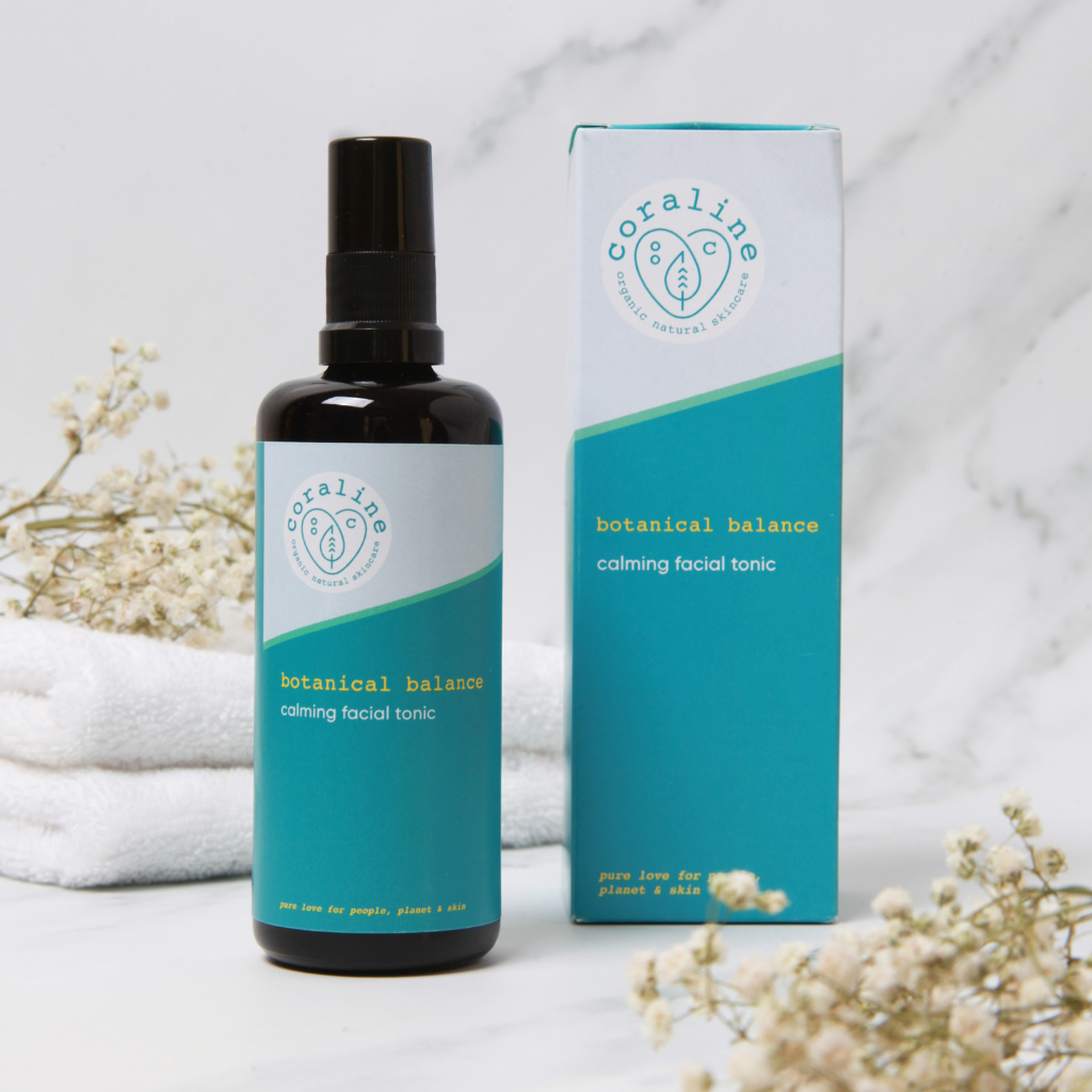 coraline skincare botanical balance calming facial tonic is in a black bottle with a blue label and is next to the outer packaging in a white and grey marble background with white towels and some dried cream flowers