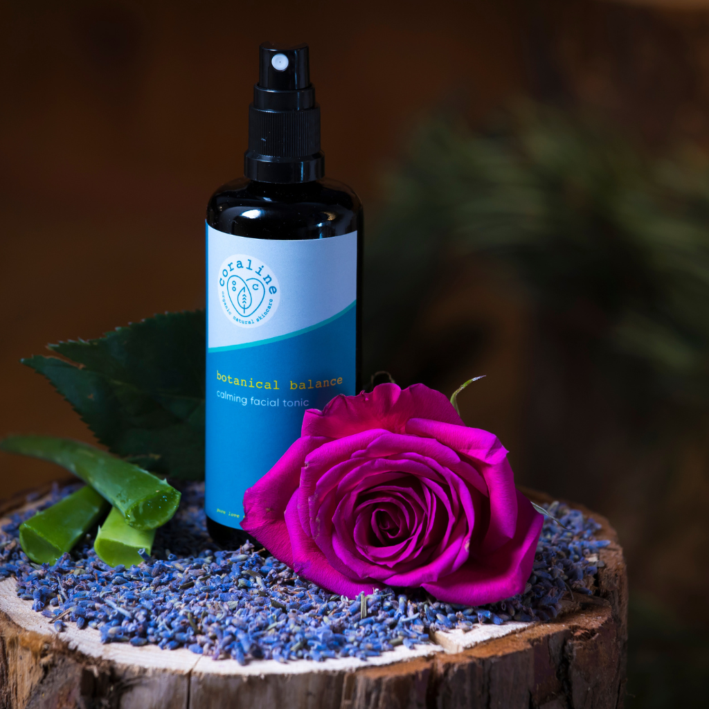 bottle of 100% natural facial tonic is sitting on top of a tree log with fresh ingredients that are inside the tonic next to it including cut aloe vera, a dark pink rose and dried blue lavender
