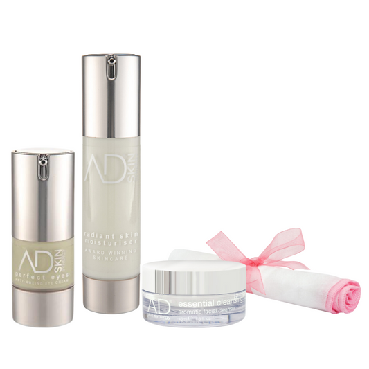 A set of the AD skin synergy products arranged against a white background. The set includes a round jar of the cleansing balm with a white and silver cap, two cylindrical pump bottles of the eye cream and moisturise with silver pumps, and a small rolled white muslin cloth tied with a pink ribbon.