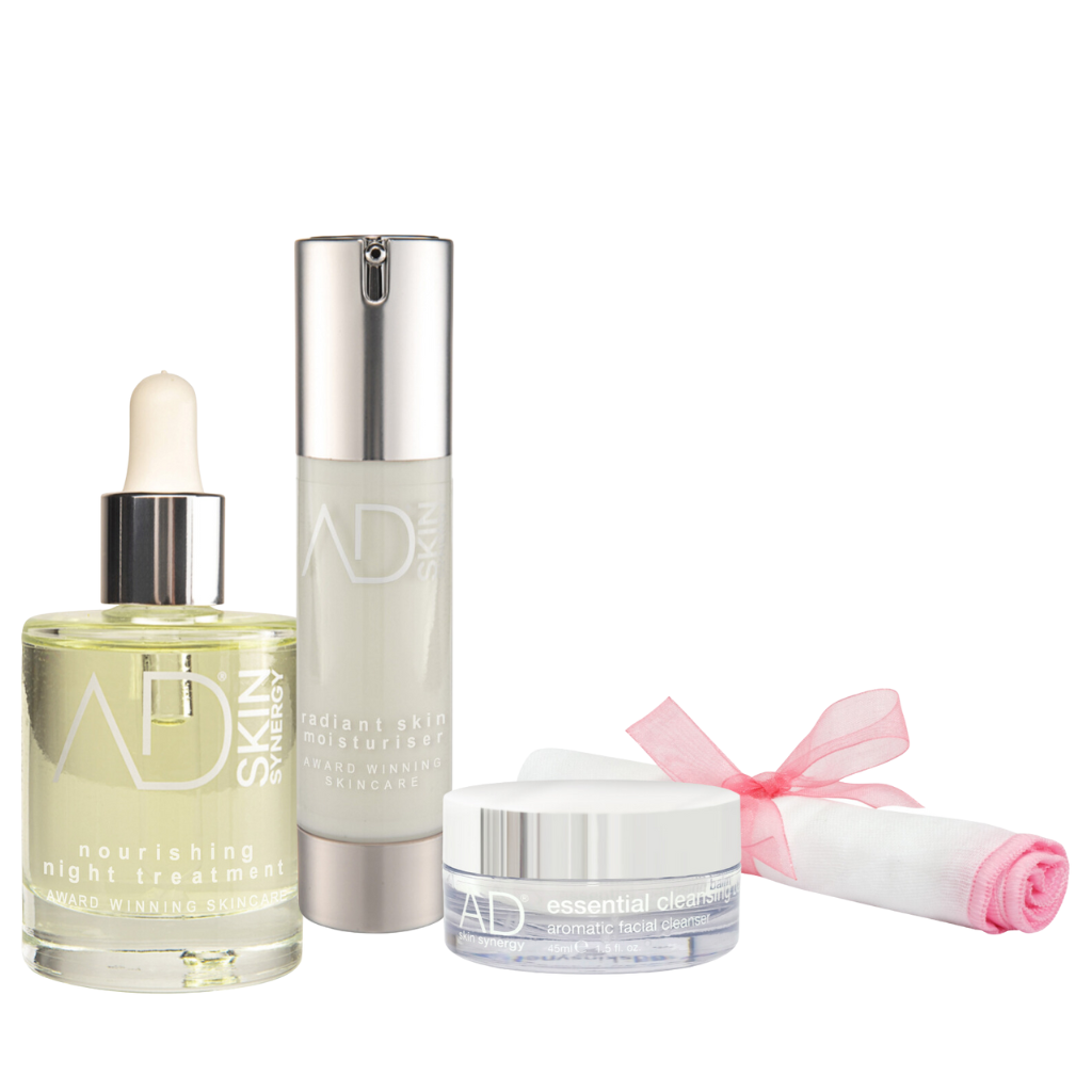 A set of the AD skin synergy products including a tall pump bottle, a jar of the cleansing balm, a dropper bottle filled with a golden liquid, and a rolled-up pink organic muslin cloth tied with a pink ribbon. All containers are labeled with the "AD SKIN" logo in silver text.