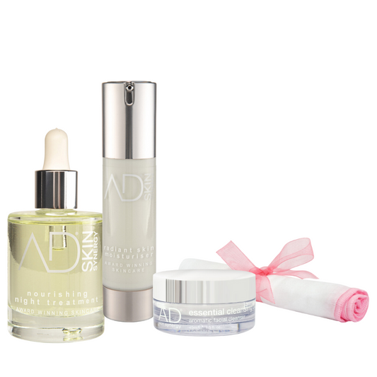 A set of the AD skin synergy products including a tall pump bottle, a jar of the cleansing balm, a dropper bottle filled with a golden liquid, and a rolled-up pink organic muslin cloth tied with a pink ribbon. All containers are labeled with the "AD SKIN" logo in silver text.