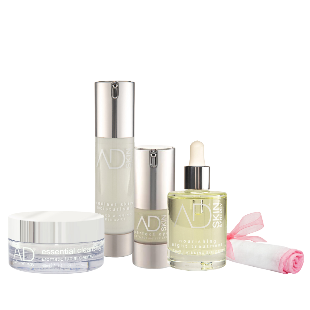 A set of the AD skin synergy products including a 2 pump bottles (with the moitsuriser and eye cream), a jar of the cleansing balm, a dropper bottle filled with a golden liquid, and a rolled-up pink organic muslin cloth tied with a pink ribbon. All containers are labeled with the "AD SKIN" logo in silver text.