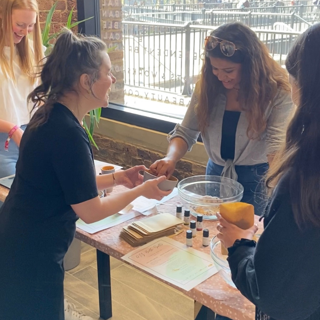 photo of 3 people enjoying a workshop on how to make your own bath salts in london. You can see a table with bowls of salts and bottles of essential oils being taught by a lady in a black dress with brown hair in a pony tail