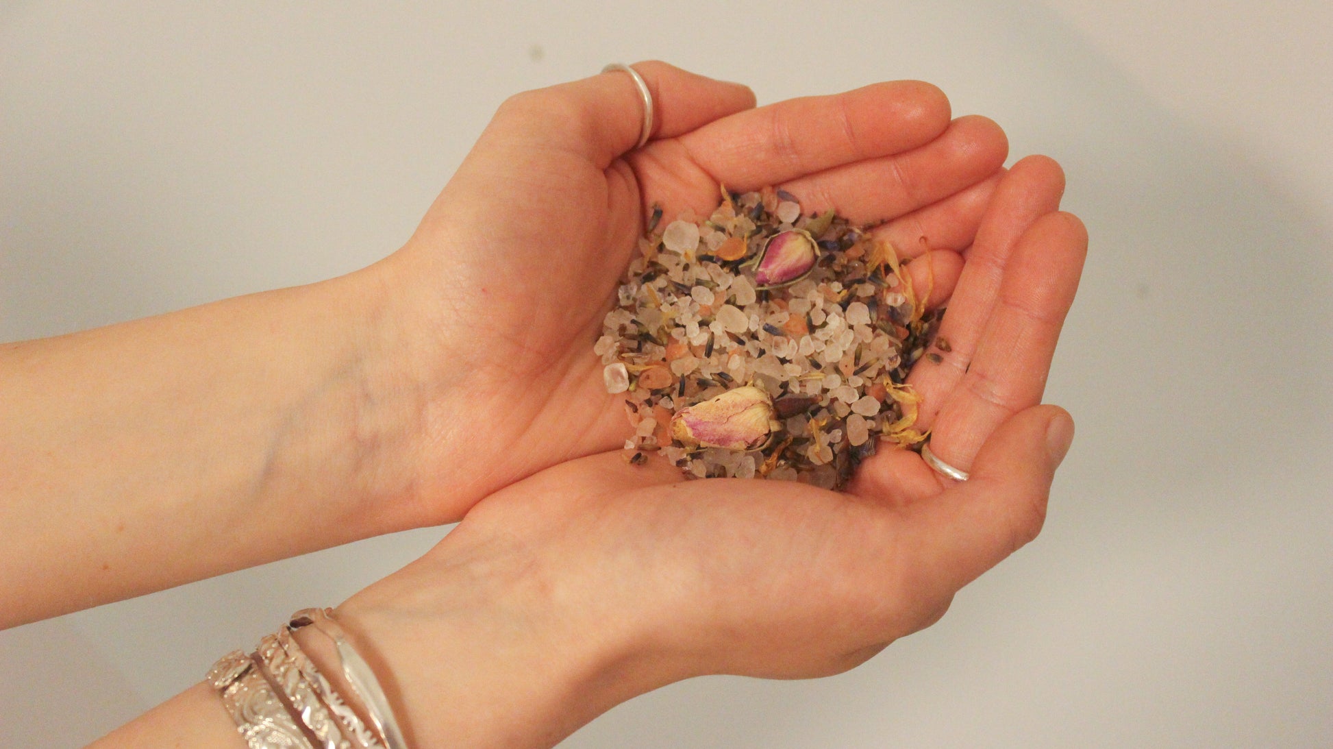 image of a white woman's hands cupped and holding some inlight beauty bath salts and wearing silver bangles and rings on their wrists and hands