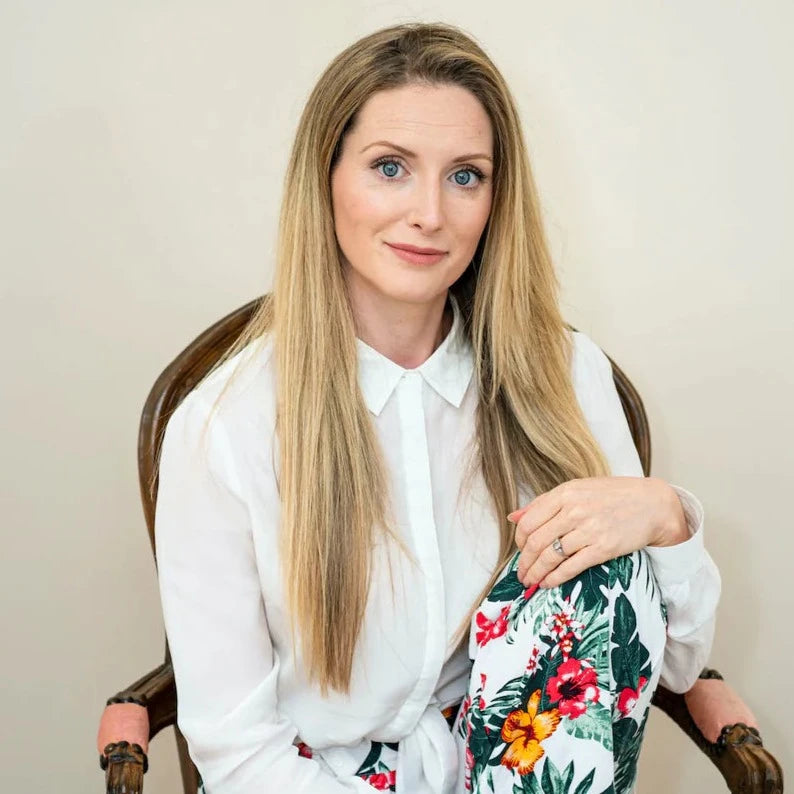 diane bowe is photographed seated on a wooden chair with dusky pink cushion. Diane is wearing a white shirt and white tropical floral pattern trousers. Diane is a hair dresser, trichologist and haircare formulator