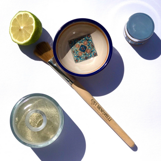 Pictured is the casa mencarelli mask mixing bowl with the mask mixing brush laid next to it. In the top left corner sits half a lime, in the top right corner is a pot of casa mencarelli balm and the bottom left corner is a bottle of the clay mask