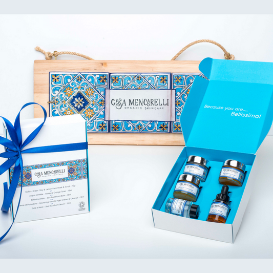 The Casa Mencarelli gift set, pictured on the left is the white gift box as it looks closed adorned with a blue bow and to the right in the box open showing the 5 products included showing a sea blue colour inside. Behind lays classic Umbrian tiling on wood with the Casa Mencarelli name and logo
