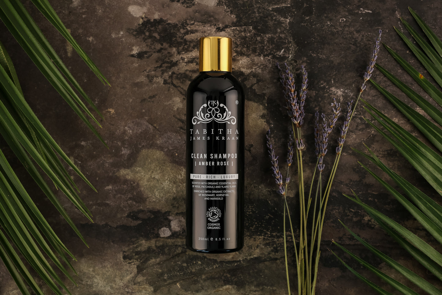 black and gold bottle of organic shampoo on a dark grey mottled marble surface with ferns and lavender fronds laid next to the bottle