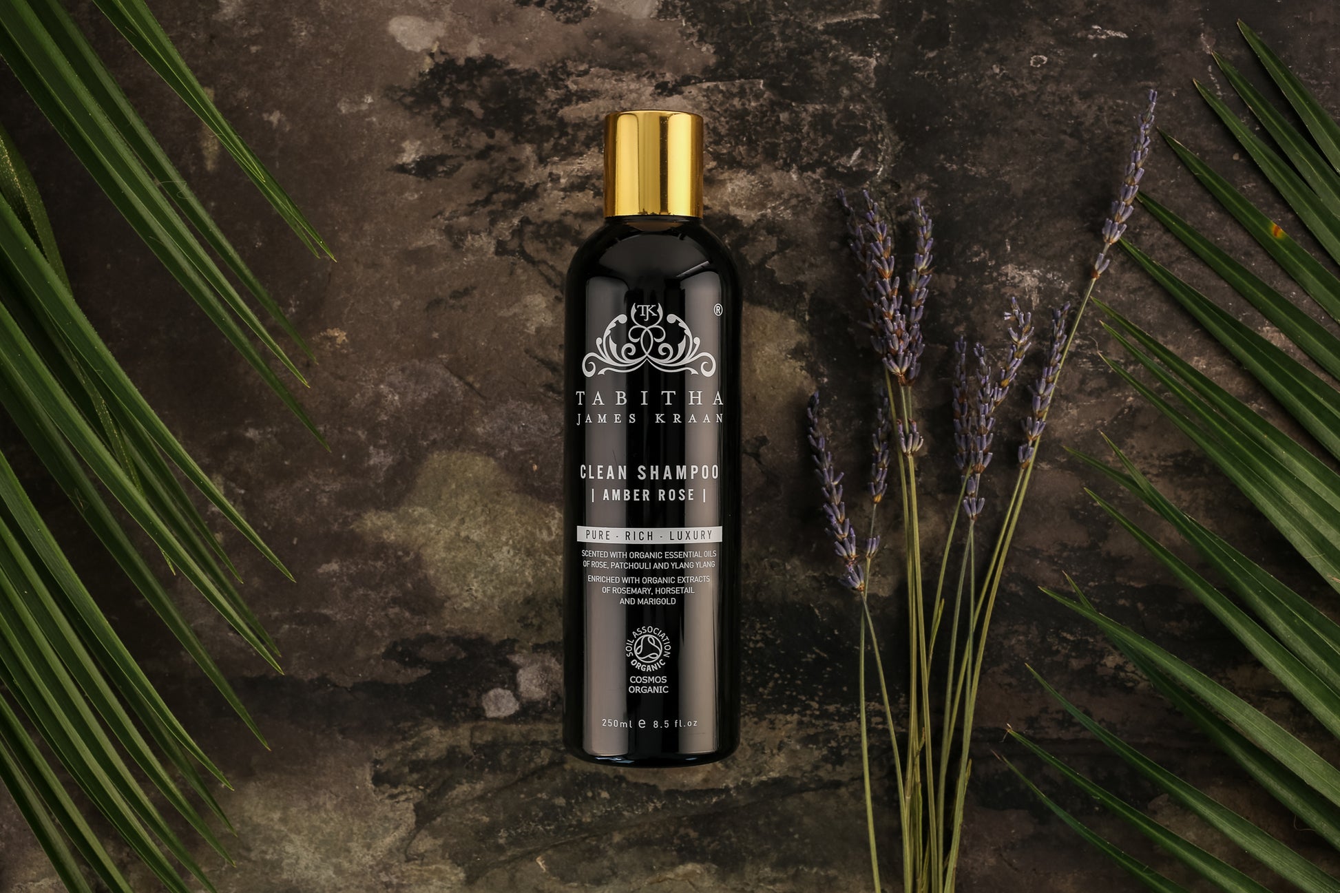 black and gold bottle of organic shampoo on a dark grey mottled marble surface with ferns and lavender fronds laid next to the bottle