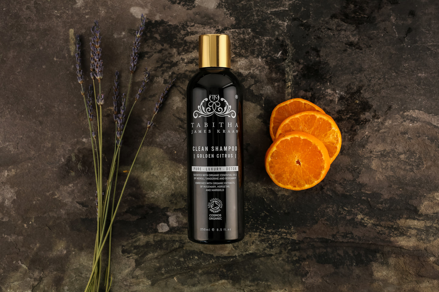 bottle of tabitha james kraan golden citrus clean shampoo. Black bottle with gold lid on a black marble background with lavender plants and orange slices laid next to it 