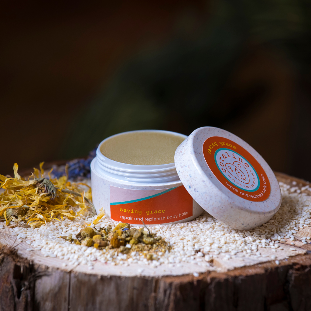 A jar of "Saving Grace" repair and replenish body balm sits open on a rustic wooden surface. The white jar with an orange label is surrounded by dried yellow and white flowers and seeds, creating a natural, earthy setting.