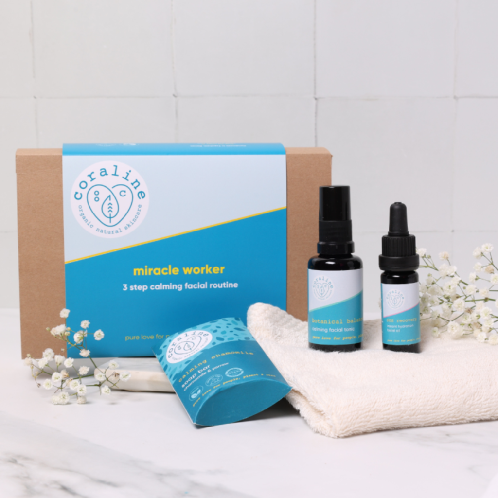 Pictured is 1 black spray bottle, 1 black dropper bottle and 1 chamomile soap. They are all placed on a soft cloth, in front of the brown Kraft box packaging and white flowers 