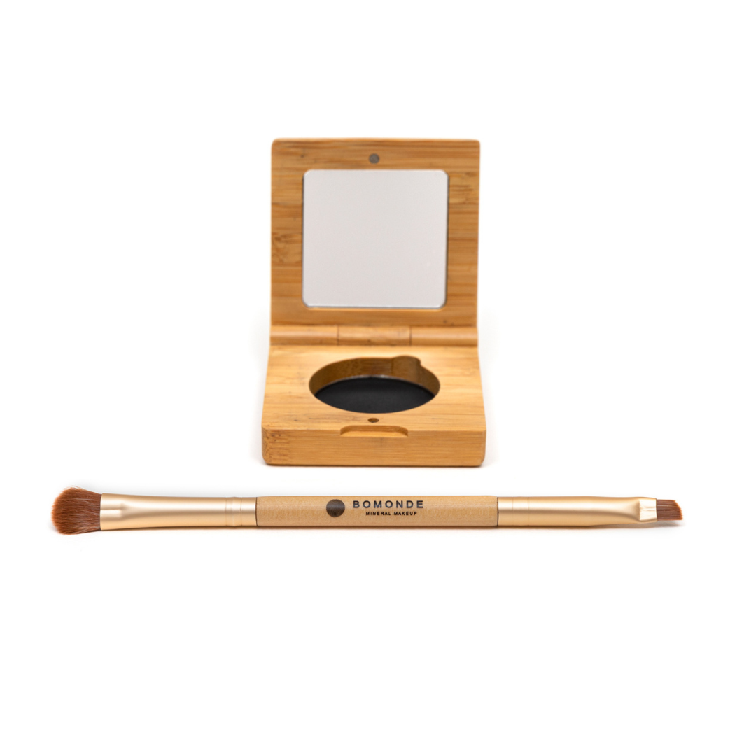 photo of bomonde mineral make up's eyeliner palette and double ended eye brush on a white background. the eyeliner is pressed into a round pan and encased in a bamboo compact case with mirror. the brow powder is dark brown. in the foreground there is a double ended eye brush made from gold metal and bamboo. the brushes are synthetic bristles. one end is a fluffy eyeshadow brush and the other end is a compact, angled brush for eyeliner and eyebrow powder