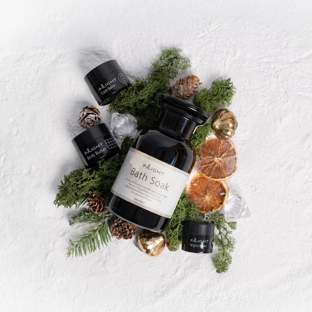 flat lay of black glass apothecary bottle of bath soak with small pots of foot balm, body butter and night balm around it on a bed of sea moss, pine needle, cone and gold baubles and dried orange slices