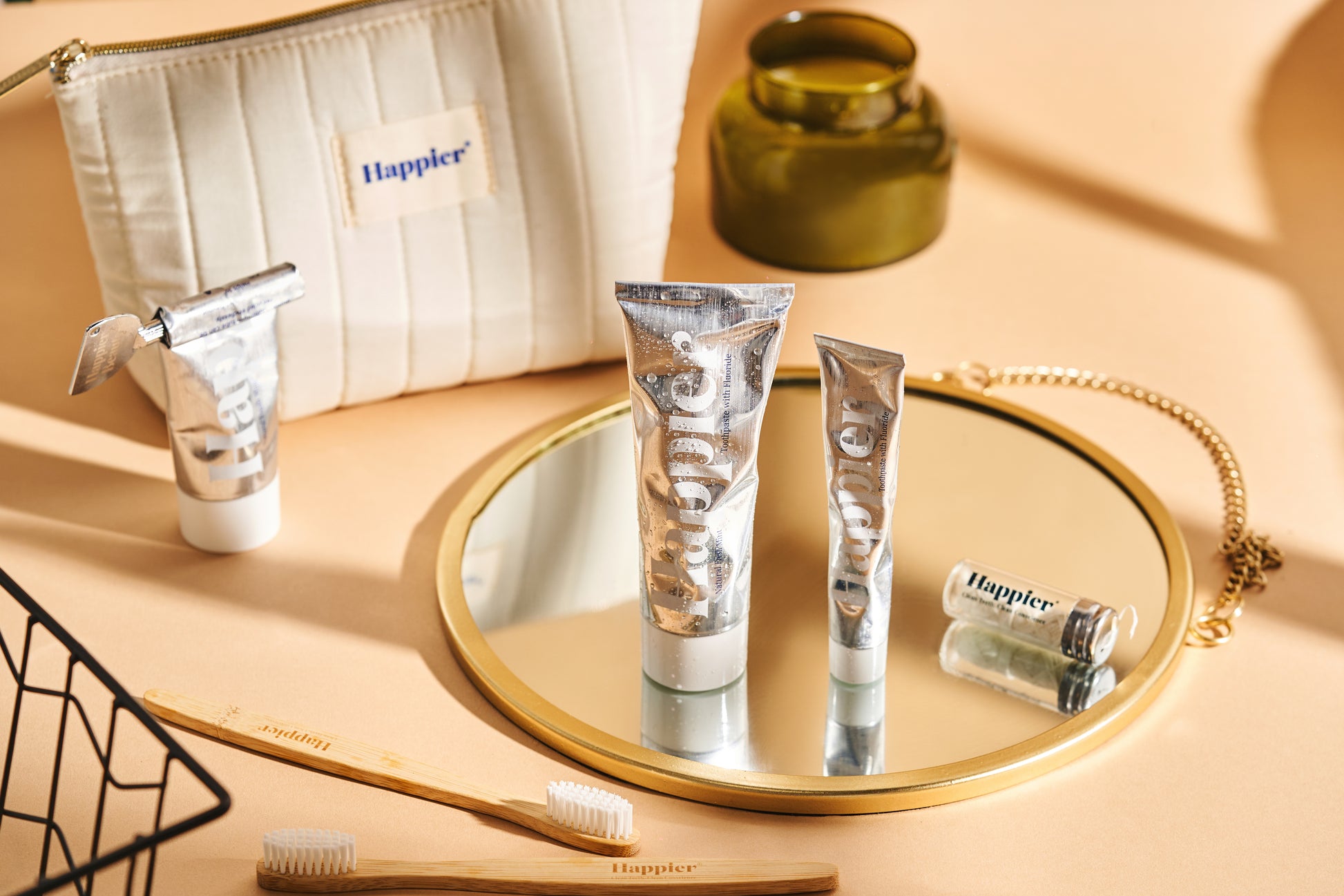 happier beauty natural tooth care products on a peach background and sitting on a round mirror with a gold/brass edge. you can see the happier beauty fabric washbag and various sizes of toothpaste tubes and the toothbrushes