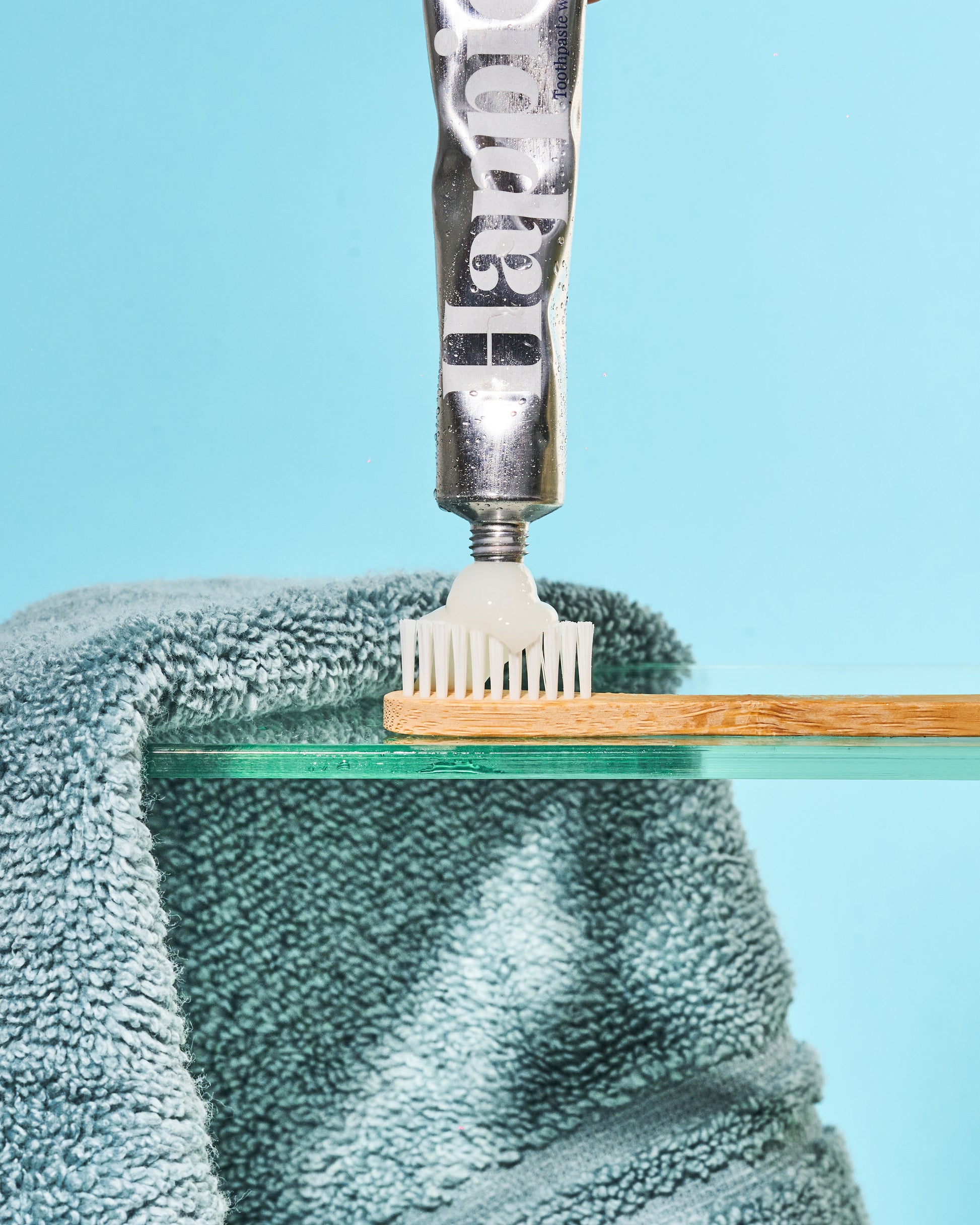 a travel sized happier beauty natural whitening toothpaste tube being squirted on to the white bristles of the bamboo toothbrush by the same brand. On a turquoise background with turquoise towel