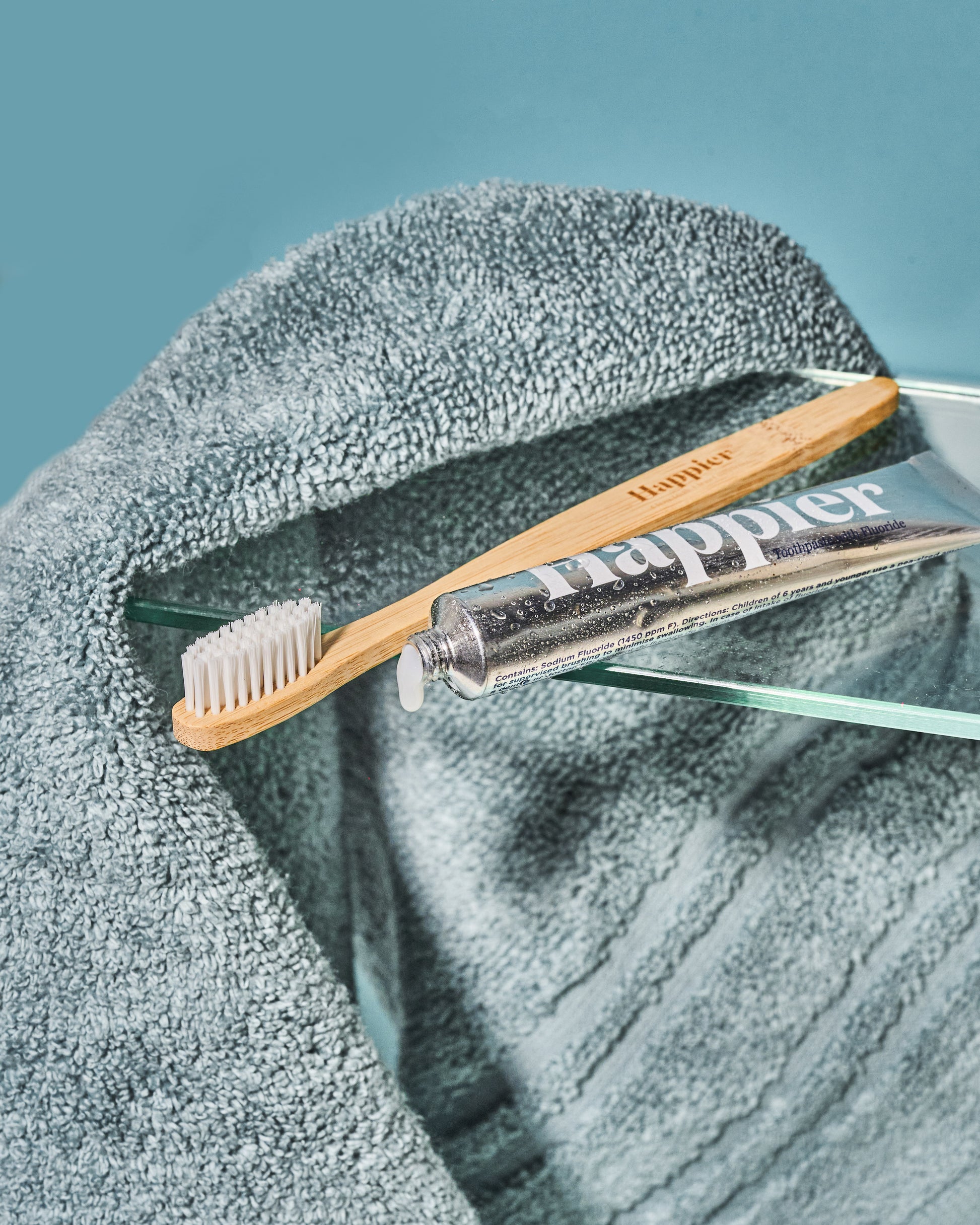 bamboo toothbrush and happier beauty natural toothpaste travel size in an aluminium tube laid flat on a clear bathroom shelf with turquoise/teal towel draped over it