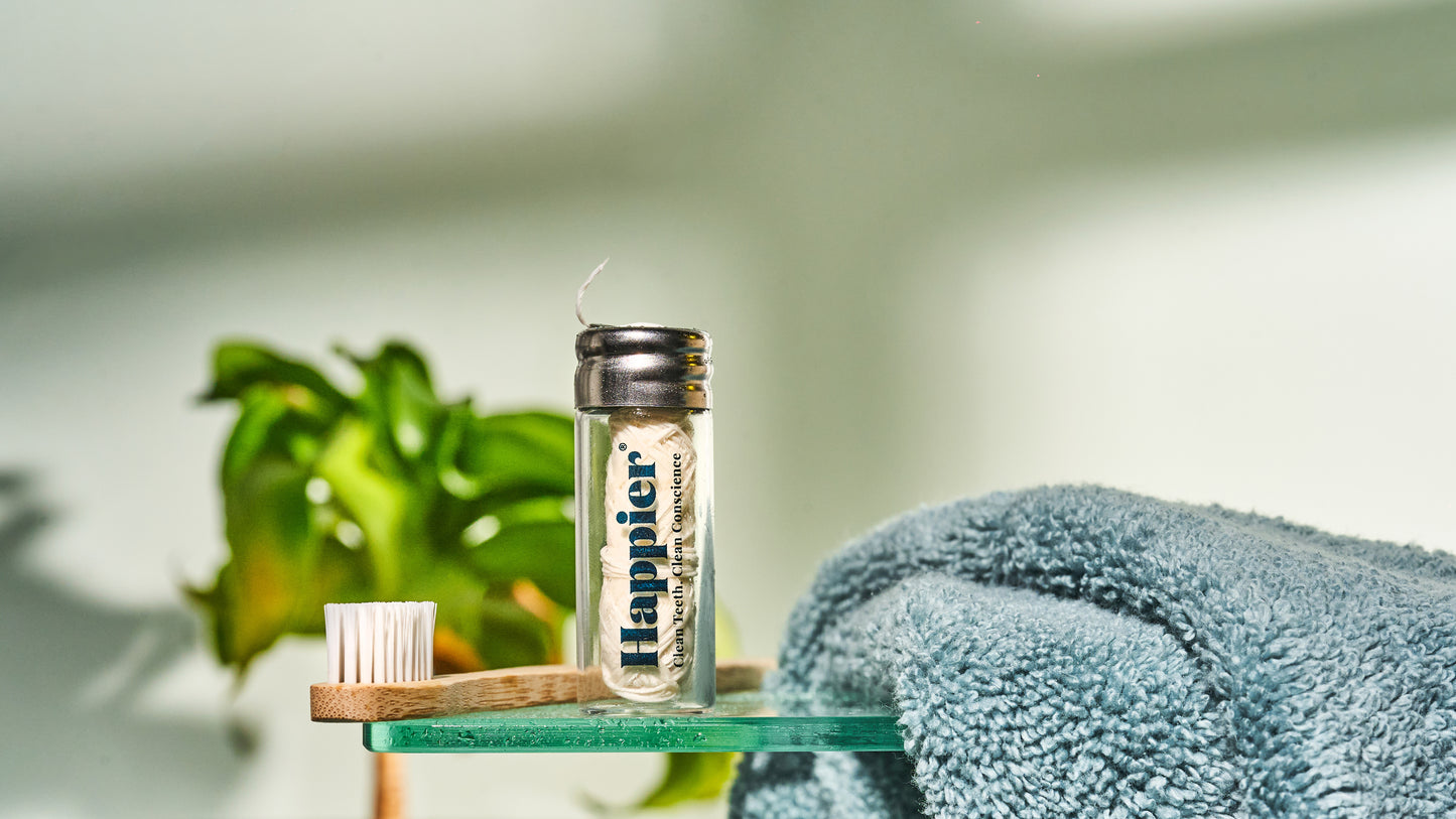 happier beauty natural dental products. toothbrush and refillable dental floss in a glass vial sitting on a glass shelf next to a light blue towel and a green plant out of focus in the background